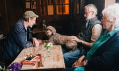 A dog sits half on a man's lap and half on a pub table amid a group of people