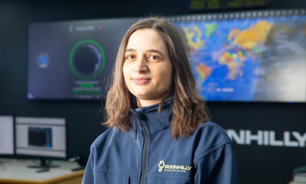Beth Sheppard wearing a Goonhilly Earth Station zip work jacket with a large screen with a world map and data and some computers in the background