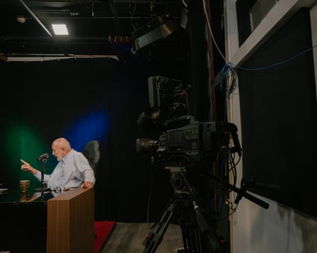 Mario Silva in a blue shirt sits behind a desk in a studio with a camera trained on him