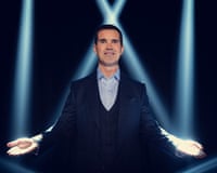 Jimmy Carr gestures with open palms, illuminated by dramatic blue stage lights