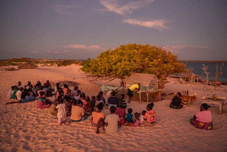 A group of about 50 people sitting on a beach at what appears to be sundown surrounding a wooden platform that has a small shrine-like shelter, into which a man is leaning. Bottles have been placed on nearby tables