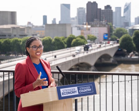 Kemi Badenoch speaks from behind a pale wooden lectern on an outdoor terrace overlooking the River Thames and a bridge. There are tall office buildings in the background. She wears a long red jacket over a bright blue top and has her braids tied back into a low bun at the back of her head.