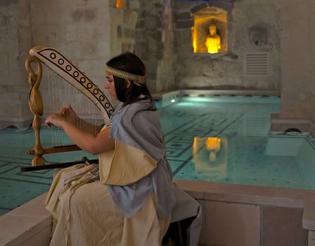A female musician dressed in traditional clothing plays a trigonon – a small triangular harp – by a shallow pool in a spa in a cave