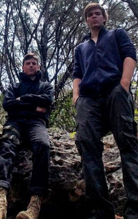 Taken from below, an image of two seemingly tall white men looking at the camera, dressed in black pants and navy tops, in a forest.