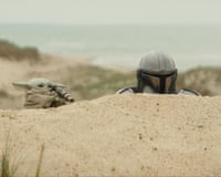 Grogu and the Mandalorian look, half concealed, over a sandy ridge.