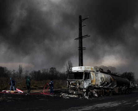 Firefighters beside a charred fuel tanker against a charred telegraph pole and smoke-filled sky