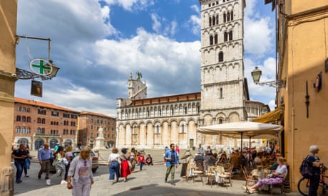 مردم در يك ميدان در لوكا، ايتاليا، با يك برج كليسا در يك روز آفتابي، و ديگران پشت ميزها زير يك چتر مينشينند.