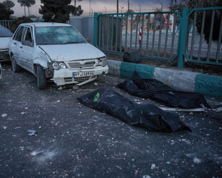 Two dead bodies in black body bags placed on the road in front of a damaged car