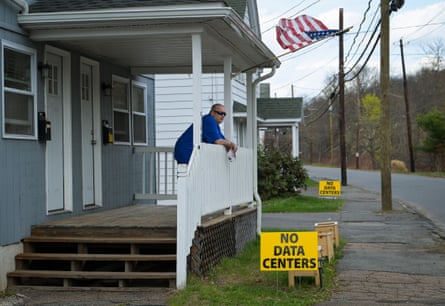 a man leaning on front porch of home, a ‘no datacenters’ sign in his lawn