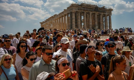 A dense crowd of tourists in sunglasses, some in baseball hats, others in vests, stand with the Acropolis in Athens in the background