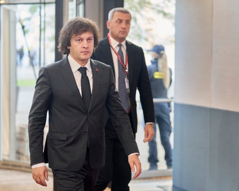 Irakli Kobakhidze walks down a corridor: he is in his late 40s with thick, dark collar-length hair and wears a dark suit; he looks burly and resolute dqxikeidqkikdinv