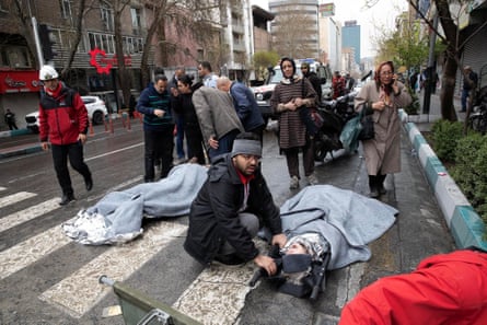 A man kneels by one of three people lying on the ground, while a crowd of distressed and concerned people looks on.