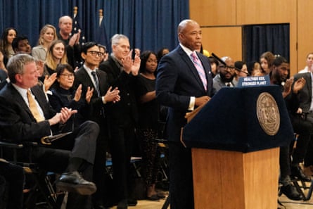 a man in a suit and tie speaks into a microphone, with a crowd of people clapping behind him