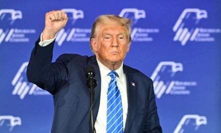 Elderly white man with orange-tinted skin and suit raises fist behind a lectern on a stage in front of a blue background.