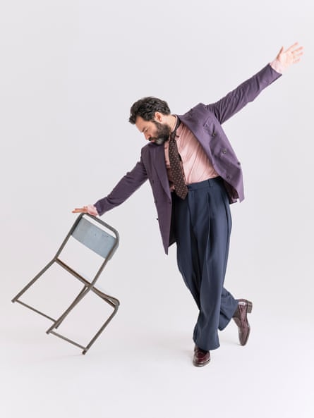 Aidan Turner, in a purple blazer, salmon shirt, brown tie with a white pattern, baggy navy trousers and shiny purple brogues, tips a chair while spreading his arms wide