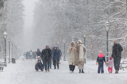 People walk along a wide, snow-covered path edged by ornate lamp-posts. Two children are sitting on a sledge in the centre.
