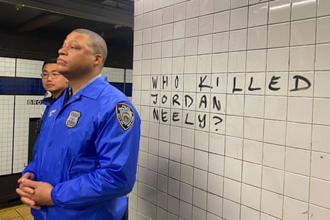 An officer stands next to a white tiled wall. On the wall, someone has written 'Who killed Jordan Neely?'