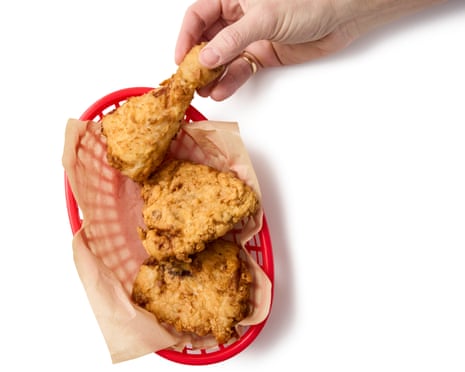 The arm, wrist and hand of a white male reaches in to a red basket containing three pieces of fried chicken, and has been successful in obtaining one of these beige snacks, for the fingers have seized the top piece by the bone end and the hand appears to be extracting the fried, battered morsel from the basket in preparation for consumption.