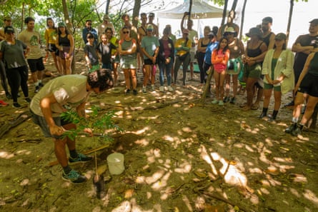A man in a yellow shirt holding a spade stands at the centre of a circle of people and prepares to plant a sapling.