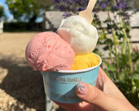 A hand holds up a blue tub containing scoops of pink, white and yellow ice-cream