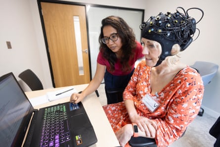 Linda Geddes wearing a cap fitted with multiple electrodes and wires sits at a desk taking part in a brain-monitoring experiment. Another woman stands beside her, pointing at a laptop screen as they review information together in a clinical research room.