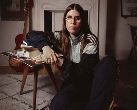 The comedian Ele McKenzie sits in a room with a small stool and a guitar in the background.