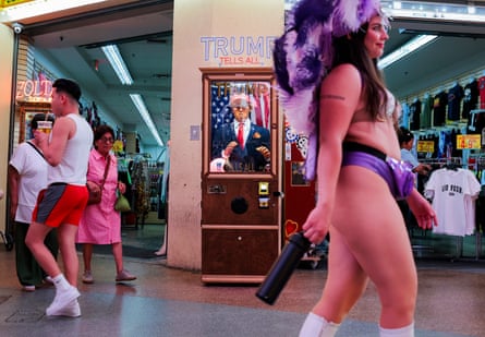 A woman walking along the street in a purple headdress, in front of a booth with a dummy of Donald Trump. A man in shorts drinking from a straw in the background. A lady in a pink dress is in the background too.