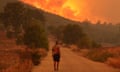 A man is seen from behind as he stands on a road looking at a wildfire. The air is smoky and the sky is orange