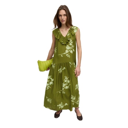 A model wears a v-neck frill detail midaxi length dress. The dress is dark green with a light green large floral print. The dress is a relaxed fit and the model is holding a lime green clutch purse.