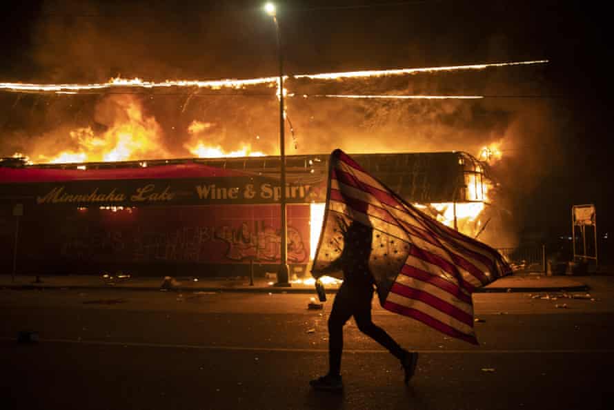 A protester carries a U.S. flag upside down, a sign of distress, next to a burning building, May 28, 2020, in Minneapolis, part of protests over the death of George Floyd