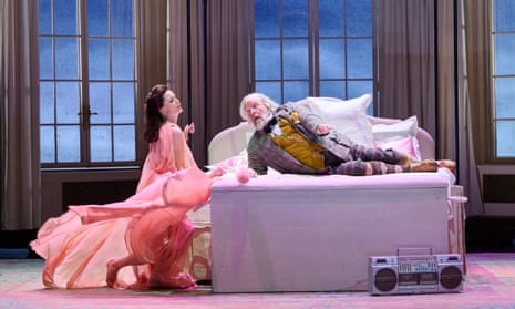 Henry Waddington as Falstaff, in yellow waistcoat, check suit and and knee-high socks, lolling on a bed, with Kate Royal’s glamorous Alice Ford, swathed in pink not quite on the bed