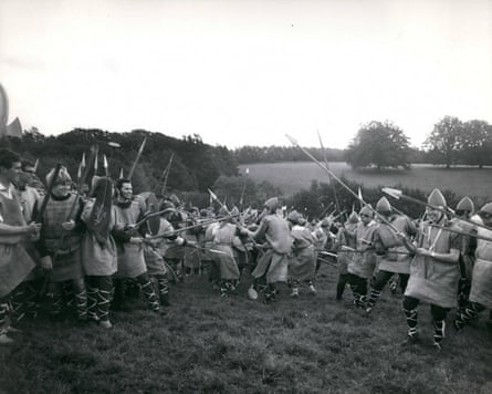 A black and white image showing a group of men in a field dressed in medieval combat wear clutching shields and raising spears