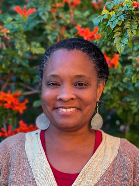 A smiling woman in round, flat earrings smiles in front of an orange and gree bush.