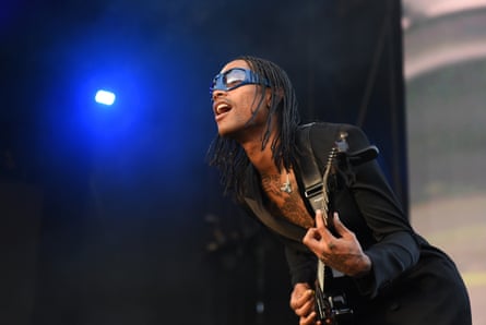Lacy dressed in dark clothing and dark glasses playing guitar