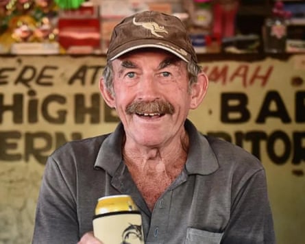 An elderly man wears a cap and proffers a beer can to the camera.