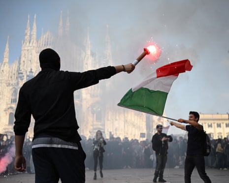 A masked protester holds a lit flare near a man waving an Italian flag outside Milan's Duomo