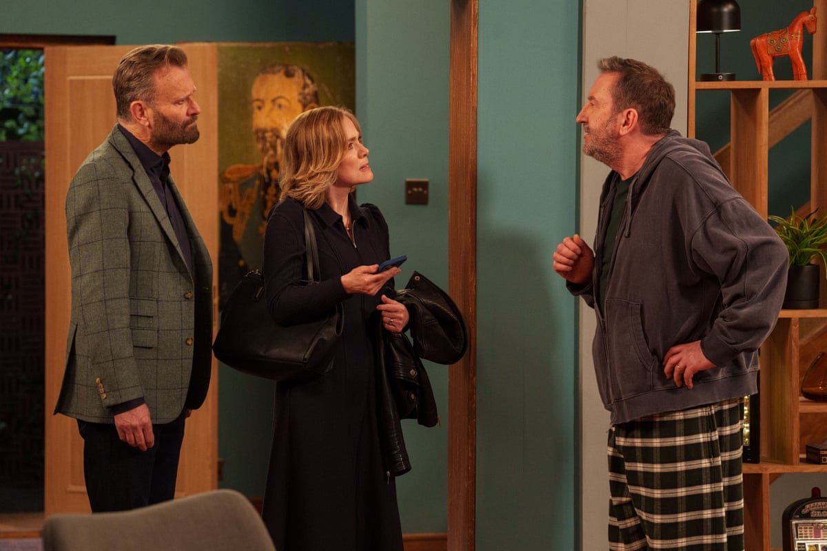 A man with a beard wearing a suit jacket and a woman in black with a jacket over her arm holding a phone speak to another man with stubble, wearing a grey hoodie and checked pyjama bottoms
