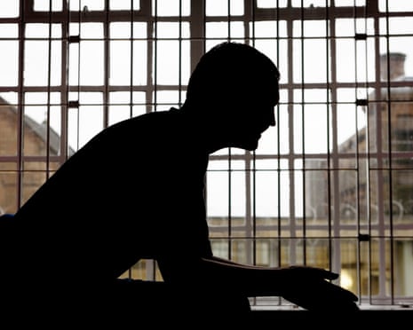 A man sits in a prison cell silhouetted by its barred window.