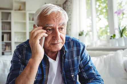 Senior man talking on mobile phone; he has white hair and sits on a white sofa in a white-painted living room; he wears a blue checked shirt over a white T-shirt and looks worried