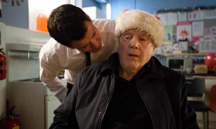 Kenneth Cope seated in a chair wearing a Santa hat, with Gray O’Brien leaning over him menacingly