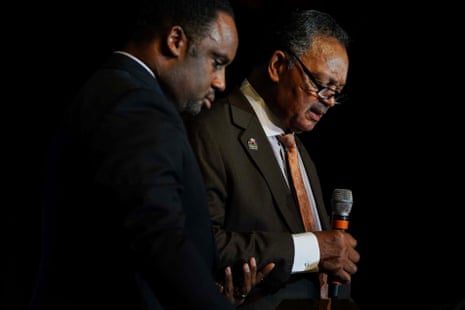 Two Black men, heads bowed, stand beside one another in suits on what appears to be a darkened stage where they are in the spotlight, with the elder speaking into a microphone he holds in one hand.