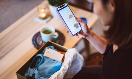 A woman looks at an online shop on her phone as she sits before a coffee table with an open package on it