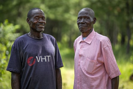 Two men, one in a blue T-shirt, the other in a pink button-up shirt stand close together outside, smiling at the camera.