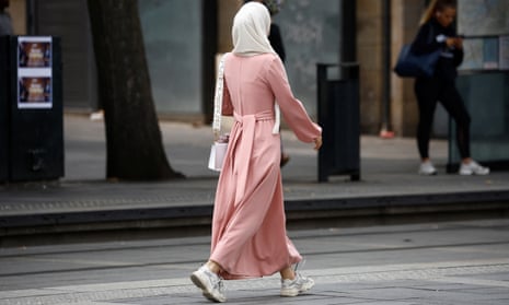 A woman walking down a street with her back to the camera, wearing wearing a pink abaya, white headscarf and white shoes.