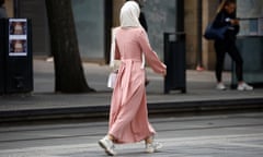 A woman walking down a street with her back to the camera, wearing wearing a pink abaya, white headscarf and white shoes.