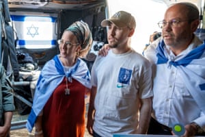 Two people draped in the flag of Israel put their arms around the shoulders of Eitan Mor, who is wearing a white T-shirt and light tan-coloured New York Yankees baseball cap