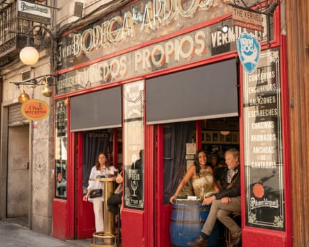 Customers sitting on barstools next to barrel tables at the open doorways of a red-painted bar with signage saying ‘Bodega de la Ardosa’