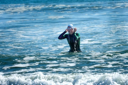 A woman in a wetsuit and swim cap pulls at her goggle, in ocean water up to her waist.