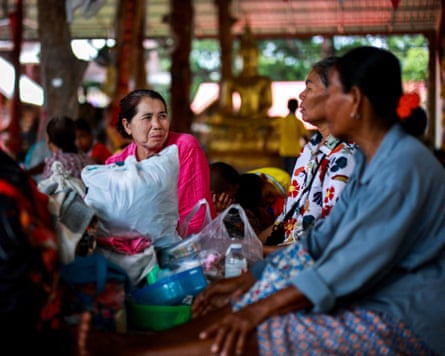 Three women sit inside an open-sided building which looks like a temple; there is a gold Buddha statue in the background. They sit among bags of possessions and provisions.