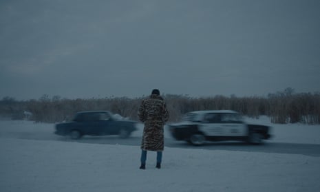 A person standing in the snow watching two cars drive in a scene from the film Tolyatti Adrift